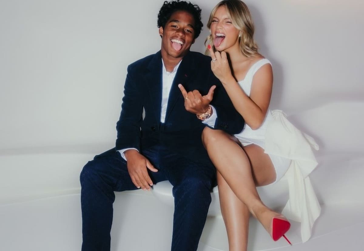 Playful couple sitting on a white couch, sticking out their tongues and smiling; man in a navy suit, woman in a white dress with red heels.