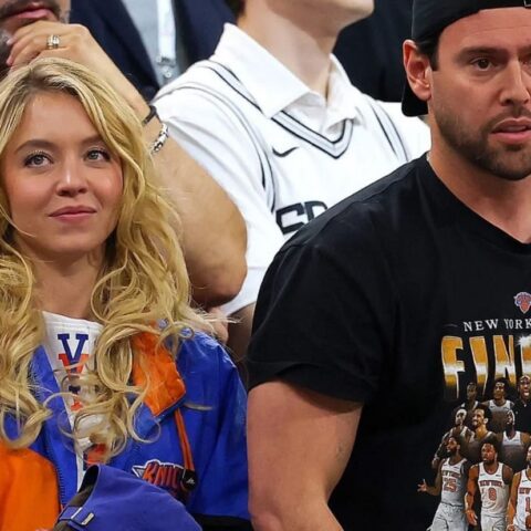Blonde woman in orange-blue jacket sits in a crowd beside a man wearing a Knicks Finals t-shirt, both watching the game steadily.