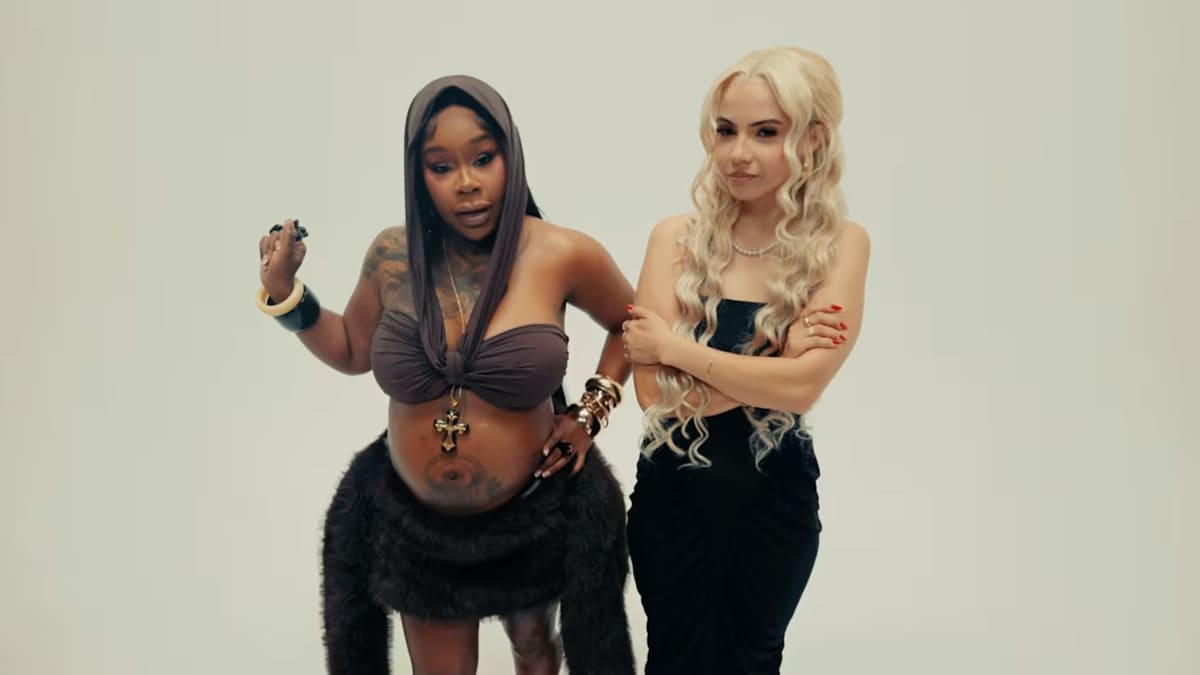 Two women pose together in a studio: one with a baby bump wearing a brown halter top and furry skirt, the other in a black dress with long blonde hair.