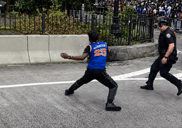 Man in a blue basketball jersey with 'Robinson 23' runs across a street as a police officer pursues behind him, with a crowd behind barricades.