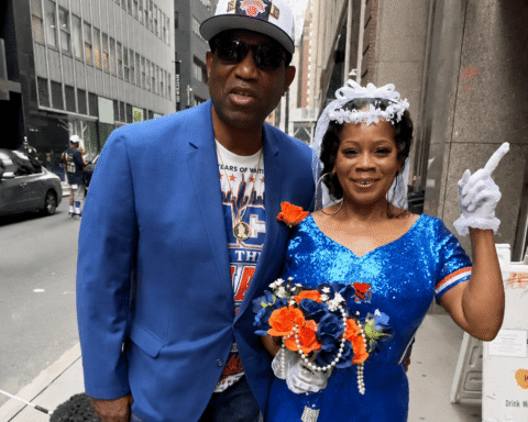 Couple posing on a city street; man in a blue blazer and cap, woman in a sparkly blue dress with a veil holding a bouquet and raising a gloved finger.