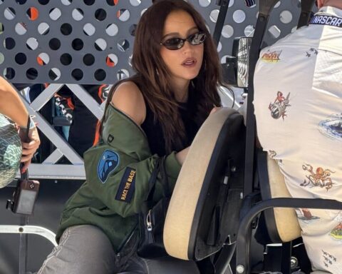 Woman wearing sunglasses and a green jacket with patches sits in a vehicle cockpit, talking to a crew member behind a perforated metal barrier.
