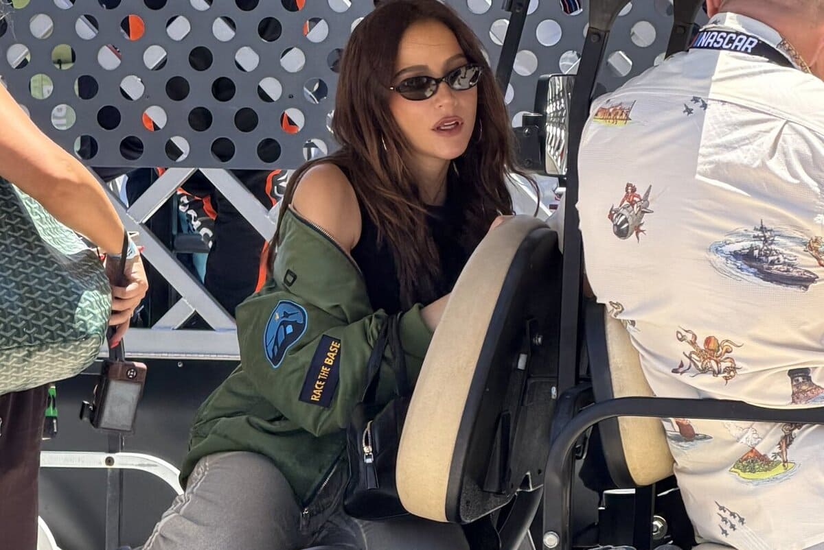 Woman wearing sunglasses and a green jacket with patches sits in a vehicle cockpit, talking to a crew member behind a perforated metal barrier.