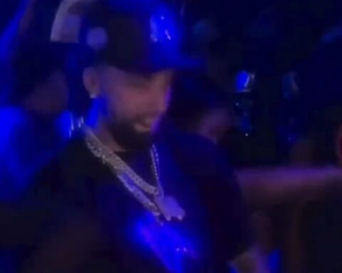 Man in a dark club under blue lights, wearing a cap and a chunky chain necklace.