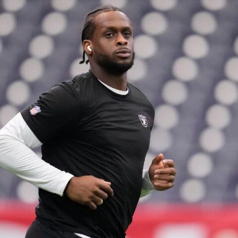 Athlete in a black training shirt sprinting on a track with white-sleeved undershirt visible and blurred stadium seating in the background.