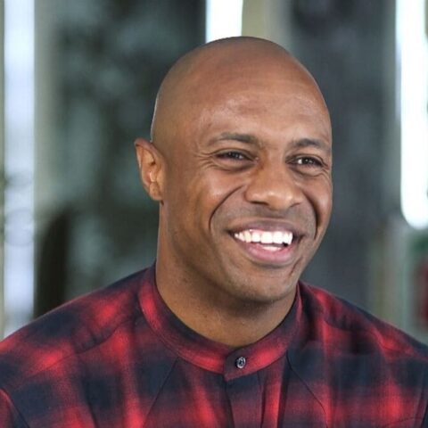 Smiling man with a shaved head wearing a red plaid shirt, indoors with a blurred background.
