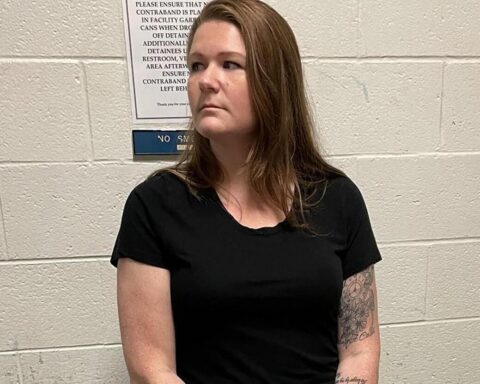 Woman with shoulder-length brown hair in a black t-shirt stands against a white cinder block wall, left forearm tattoo visible.