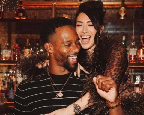 A man and woman laughing together at a bar; man wears a black striped sweater and gold necklace.