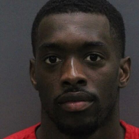 Mugshot of a young Black man with short hair, wearing a red shirt, facing the camera against a gray background.