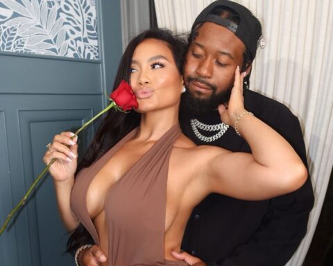 Woman in a taupe halter dress poses with a red rose near her lips while a man hugs her from behind.