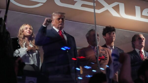 A man in a dark suit with a red tie stands center on a stage, flanked by four others including a blonde woman on his left.