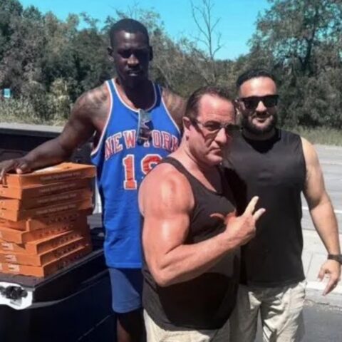 Three muscular men pose beside a pickup truck loaded with orange pizza boxes on a sunny day, outdoors by a road.
