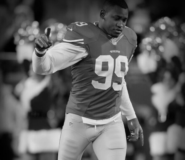 NFL player in a 49ers uniform (#99) raises his right index finger in celebration on the field (black-and-white photo).