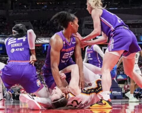 Female basketball players in purple jerseys attend to a teammate lying on the court during a game in progress to the left.