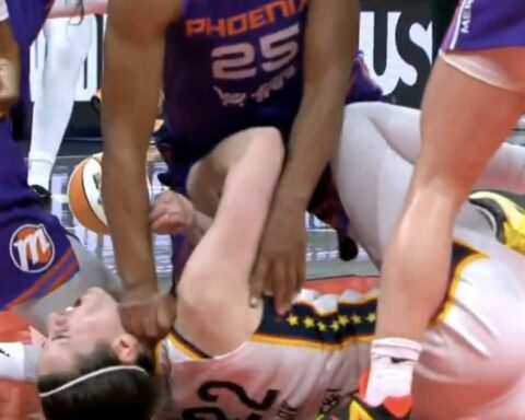 Basketball players dive and pile on the floor during a paused moment, with one player lying on his back and teammates reaching for the ball in a tumble under the basket.