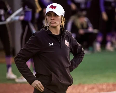 Female athlete wearing a GW white cap and black jacket stands with hands on hips on a softball/baseball field, looking to the side.