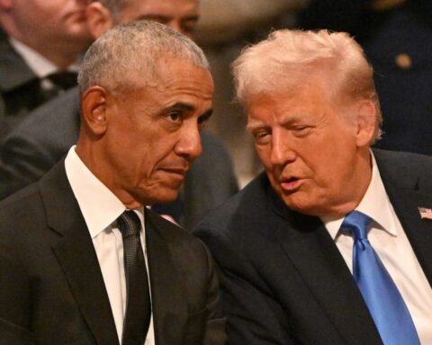 Two men in dark suits lean in for a private conversation at a formal event; one has a black tie, the other a blue tie with a flag pin.