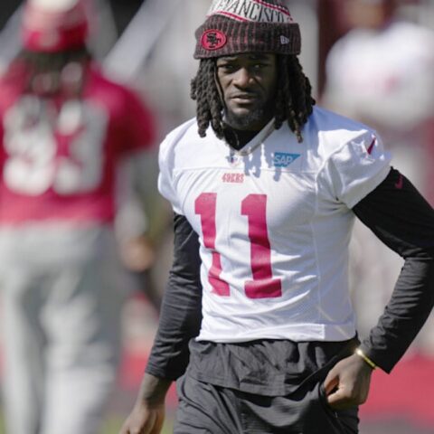 Football player in white 49ers jersey number 11 walking on the field wearing a red beanie and black sleeves.