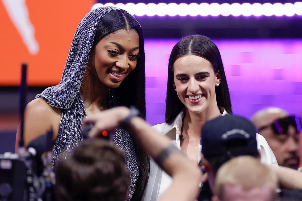 Two women pose for photographers at an event; one wears a sparkling hooded outfit, the other a white shirt, with fans and cameras in the foreground.