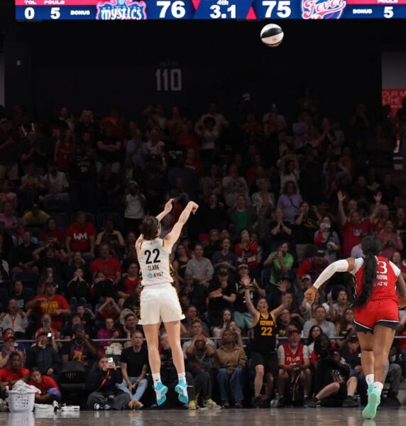 Basketball player in white jersey #22 leaps for a jump shot as the ball sails through the air, with a crowded arena in the background.