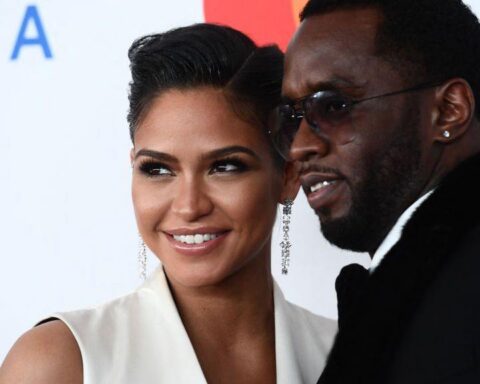 Smiling woman in a white sleeveless dress with chandelier earrings posing with a man in a black suit at a red carpet event before a light backdrop.