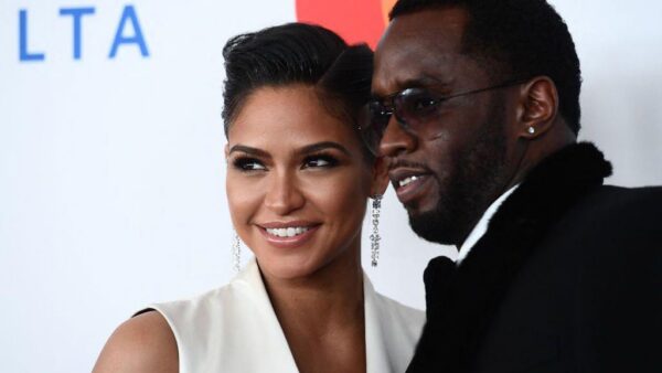 Smiling woman in a white sleeveless dress with chandelier earrings posing with a man in a black suit at a red carpet event before a light backdrop.