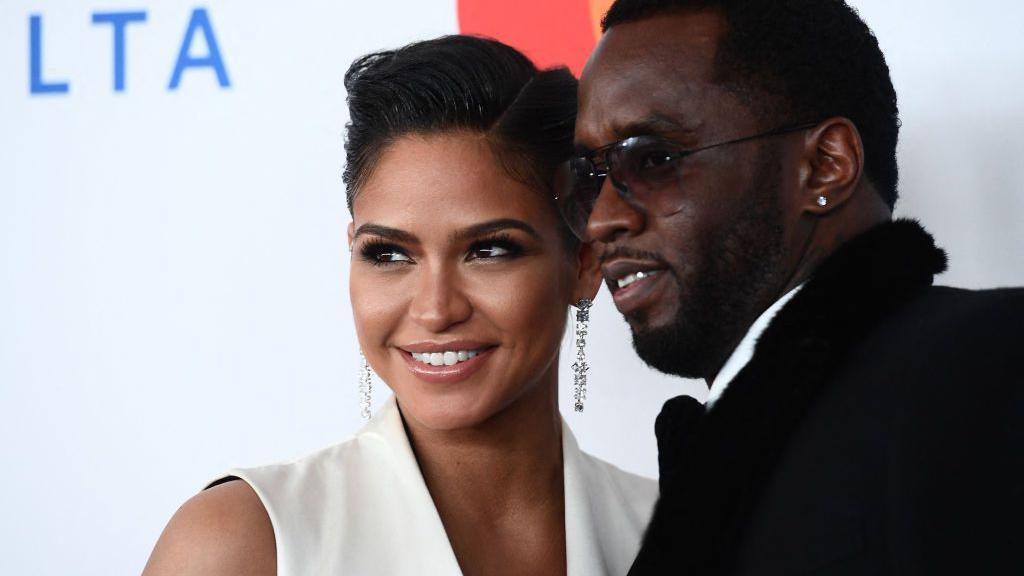 Smiling woman in a white sleeveless dress with chandelier earrings posing with a man in a black suit at a red carpet event before a light backdrop.