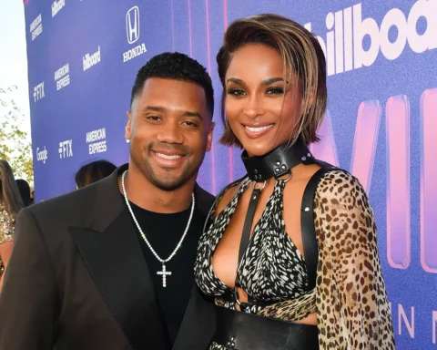 Smiling couple posing together on a purple Billboard backdrop at a red-carpet event; man in a black suit with a pearl necklace, woman in a leopard-print outfit with a black choker and cutout top.