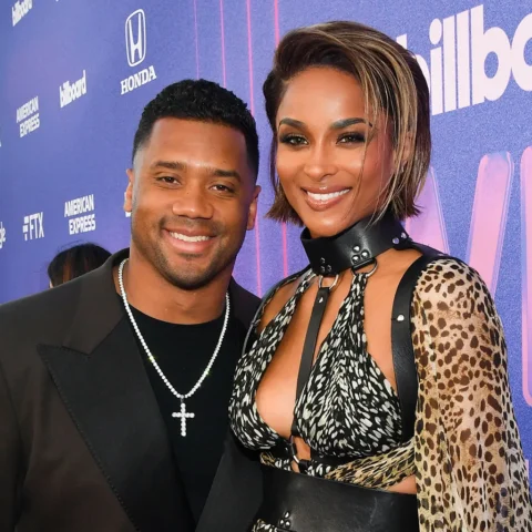 Smiling couple posing together on a purple Billboard backdrop at a red-carpet event; man in a black suit with a pearl necklace, woman in a leopard-print outfit with a black choker and cutout top.