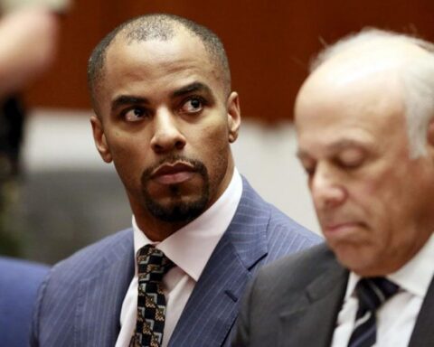 Man in a blue pinstripe suit glances to his left as another man in a dark suit sits nearby in a formal setting, likely a courtroom or meeting room.