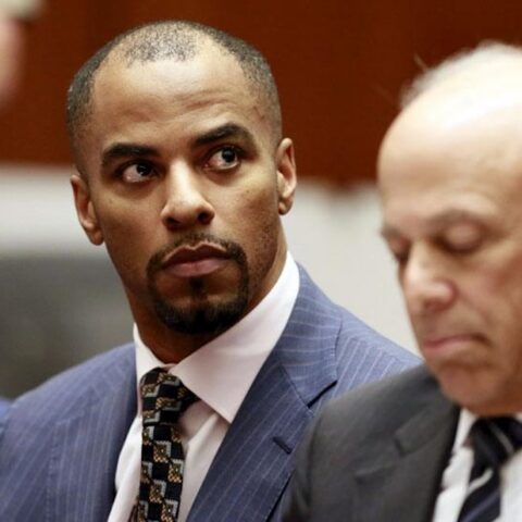 Man in a blue pinstripe suit glances to his left as another man in a dark suit sits nearby in a formal setting, likely a courtroom or meeting room.