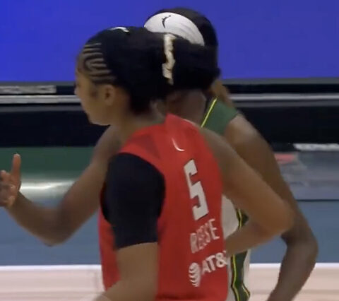Two basketball players on an indoor court; red jersey with number 5 gestures with a thumbs up while a teammate in green stands behind them.