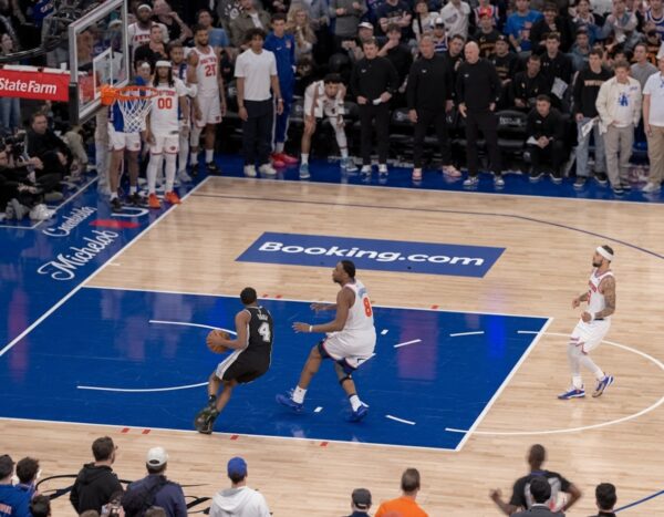 Basketball game in progress: a player in black drives with the ball as a defender in white chases, on a blue and wood court with spectators surrounding the court.