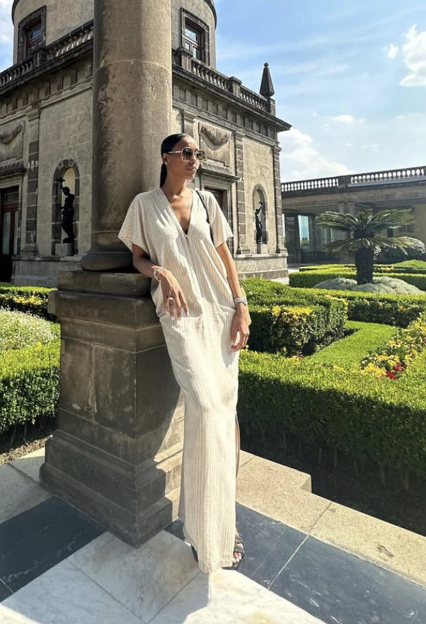 Woman in a cream striped dress leaning against a stone pillar in a formal garden with trimmed hedges and a historic building nearby.