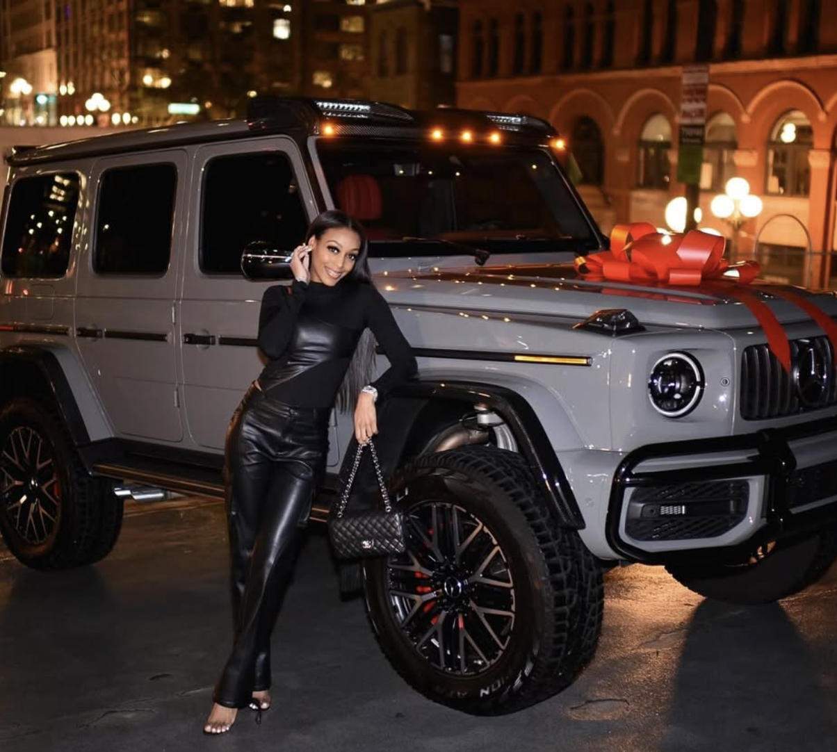Woman in a black outfit leaning on a gray Mercedes-Benz G-Class with a large red bow on the hood at night in an urban setting, holding a quilted bag.