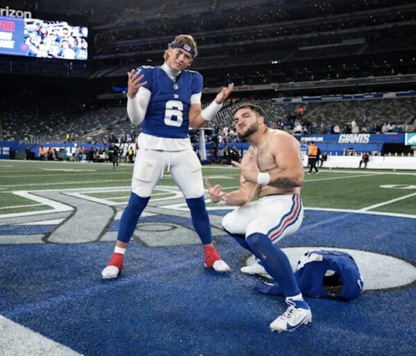 Two football players pose on the field after a game; one in blue jersey #8 stands with arms out, the other kneels shirtless with a helmet on the turf nearby.