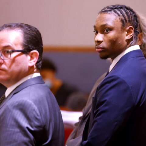 Two men in formal suits in a courtroom; one with glasses facing left, the other glancing toward the camera.