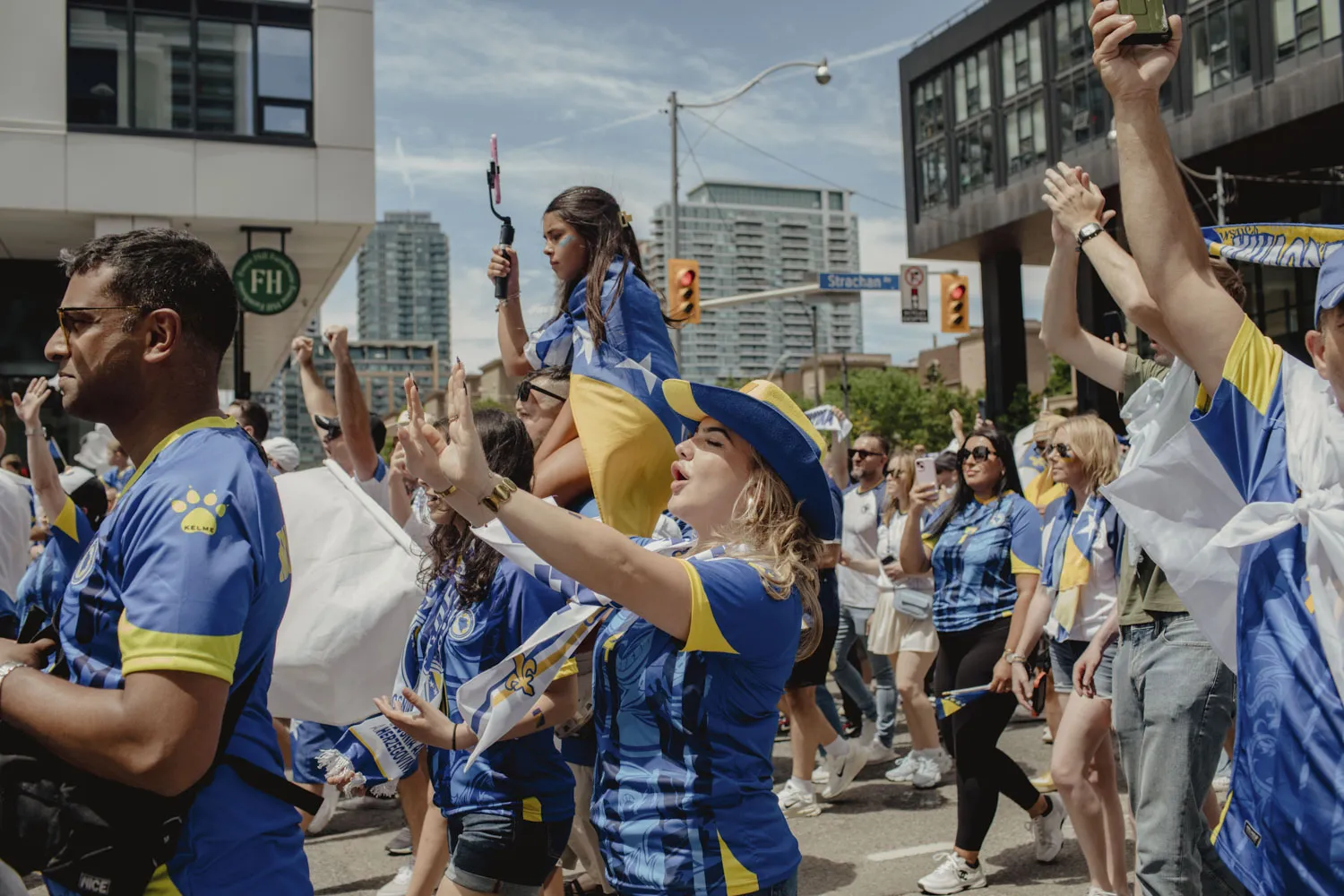 People wearing blue and yellow outfits cheer and wave flags in a city street parade; a woman in a blue hat raises her arms as others film and cheer.