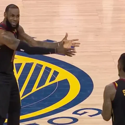 Basketball coach with arms outstretched addressing players on the court, with a player wearing a 'SMITH' jersey nearby and a large blue-yellow logo on the floor nearby.