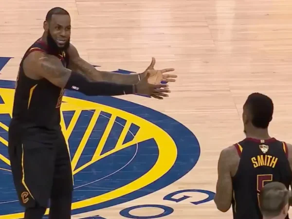 Basketball coach with arms outstretched addressing players on the court, with a player wearing a 'SMITH' jersey nearby and a large blue-yellow logo on the floor nearby.