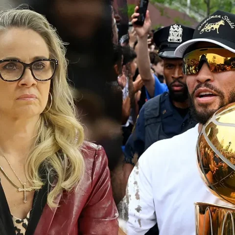 Woman with blonde hair and glasses sits with a serious expression beside a man wearing mirrored sunglasses, holding a large gold basketball trophy with a crowd behind them.