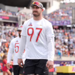 Male athlete in a white jersey with the number 97 walks on a sports field as fans watch from crowded stands behind him.