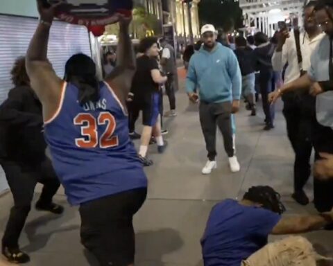 Crowd on a city sidewalk watches as a man in a blue basketball jersey with the number 32 raises a skateboard overhead, while another person kneels on the ground nearby.