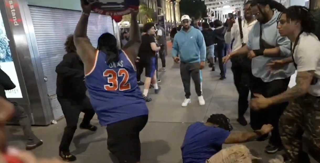 Crowd on a city sidewalk watches as a man in a blue basketball jersey with the number 32 raises a skateboard overhead, while another person kneels on the ground nearby.