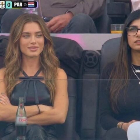 Two women sit courtside at a sports event; left woman with wavy light brown hair crosses her arms as a Coke bottle rests between their laps, scoreboard reads USA 2–0 PAR (tennis/ball sport).