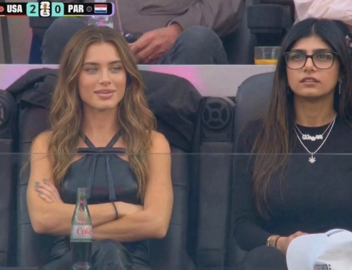 Two women sit courtside at a sports event; left woman with wavy light brown hair crosses her arms as a Coke bottle rests between their laps, scoreboard reads USA 2–0 PAR (tennis/ball sport).
