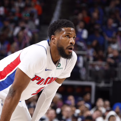 Black male basketball player in a white Pistons jersey with red and blue stripes, bent forward on the court as a crowded arena watches the game.