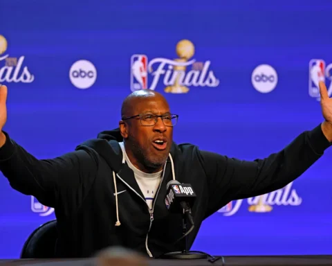 A man raises both arms while speaking into a microphone at a NBA Finals press conference backdrop.
