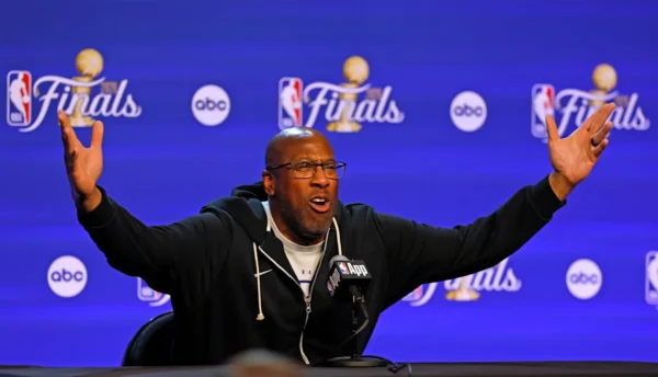 A man raises both arms while speaking into a microphone at a NBA Finals press conference backdrop.