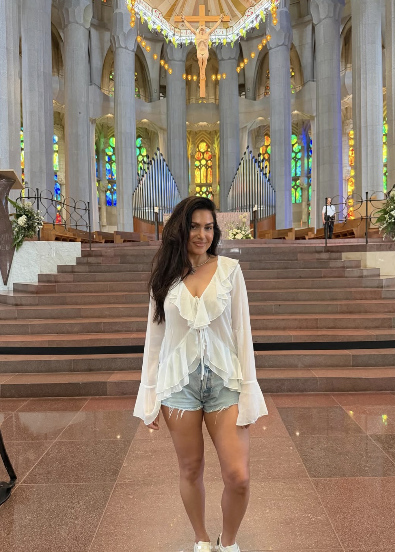 Woman posing on stone steps inside a grand church, wearing a white ruffled blouse and denim shorts with colorful stained-glass windows behind her.